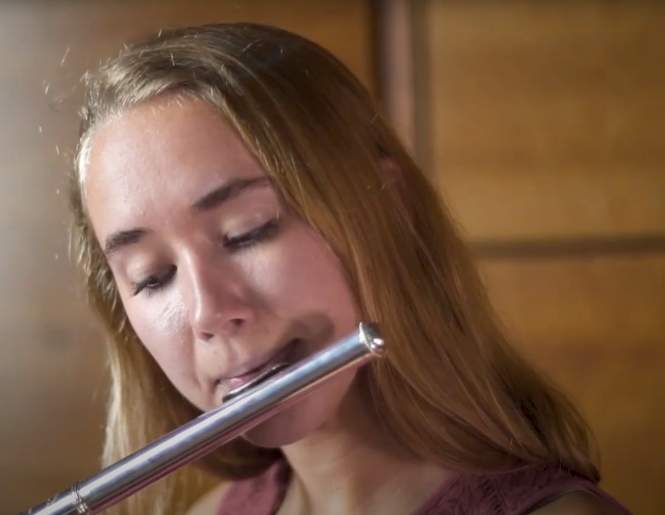 A person with long blonde hair is playing a silver flute indoors, focusing intently on the instrument.